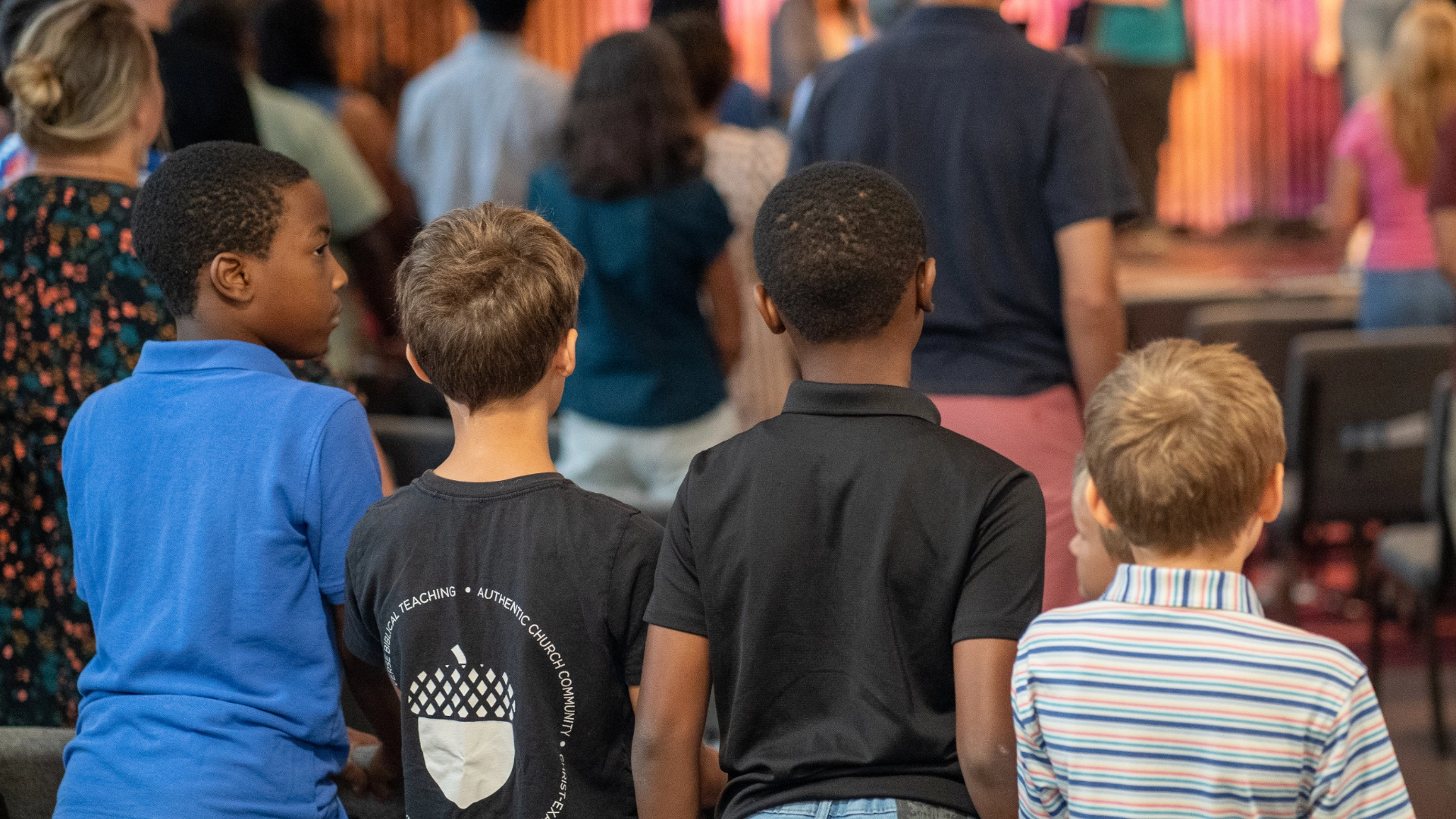 Four young boys stand in a row, facing away from the camera, in a Sunday morning church service.