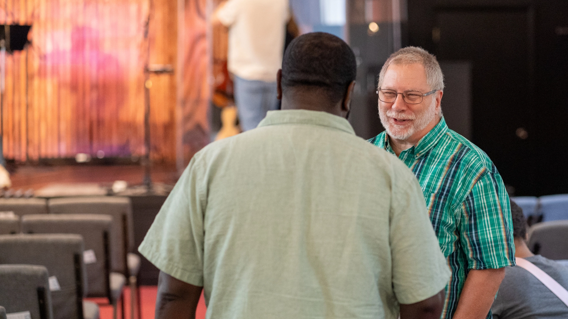 Two men are engaged in a conversation inside a room with chairs and a stage in the background. One man faces the camera while the other has his back to it.