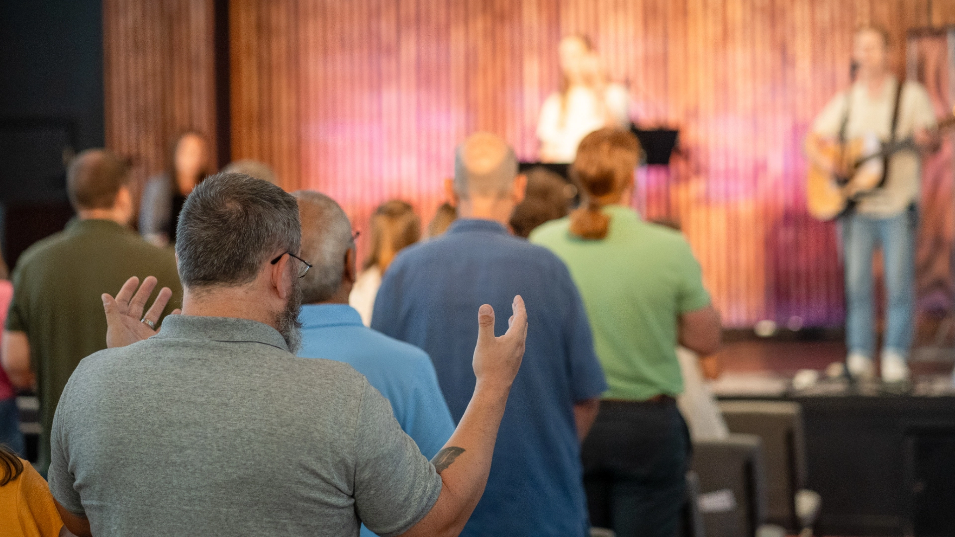 A group of people gathered at Fellowship Raleigh Church stands in a room, facing a stage where musicians play. One man in the foreground has his hands raised.